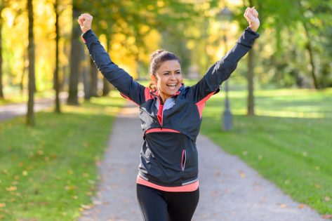 Happy fit middle aged woman cheering and celebrating as she walks along a rural lane through a leafy green park after working out jogging.