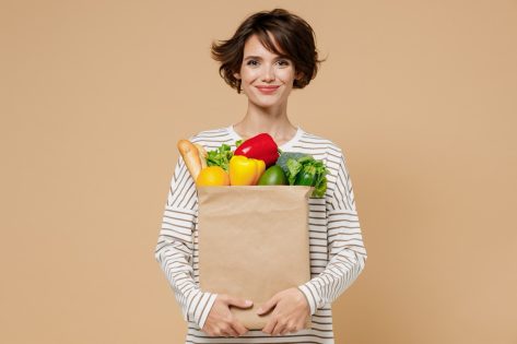 Young smiling caucasian cheerful happy fun vegetarian woman 20s in casual clothes hold paper bag with vegetables after shopping look camera isolated on plain pastel beige background studio portrait.