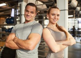 Waist up portrait of muscular couple smiling at camera while standing with arms crossed in modern gym lit by sunlight