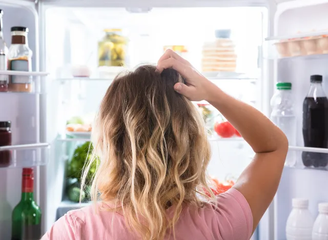 Rear View Of A Confused Woman Looking In Open Refrigerator At Home