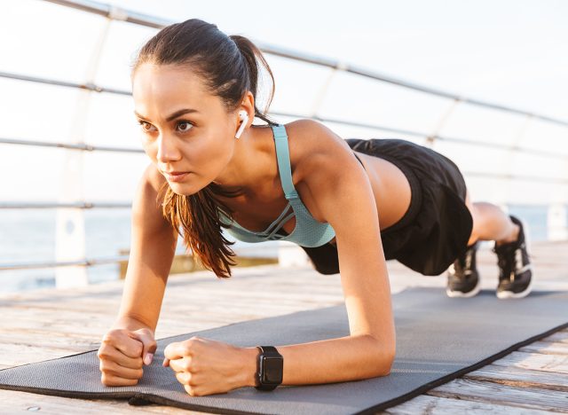 Close up of a serious asian sportswoman in earphones doing plank exercise outdoors at the beach