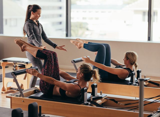 Women doing pilates exercises lying on pilates workout machines while their trainer guides them. Two fitness women being trained by a pilates instructor.
