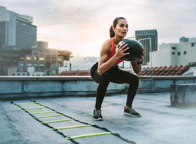 Female athlete doing squats holding a medicine ball standing on a rooftop. Woman doing workout using medicine ball with an agility ladder by her side on rooftop.