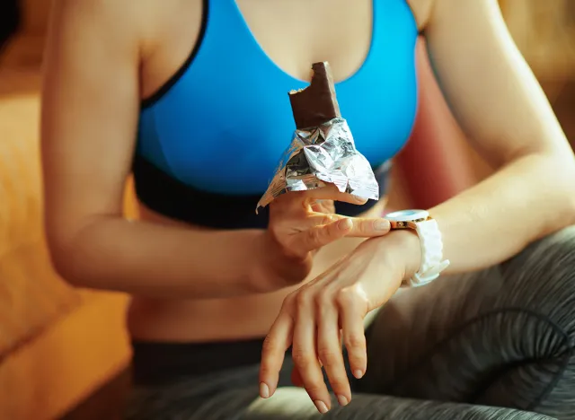 Closeup on fit sports woman in sport clothes with chocolate raw protein bar using smart watch in the modern living room.