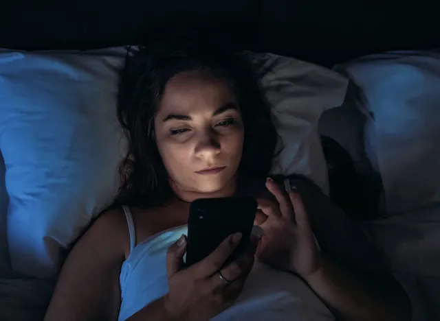 Insomnia and social media addiction concept. Young woman uses smartphone while lying in bed at night, toned