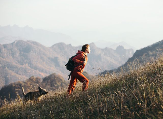 Beautiful woman traveler climbs uphill with a dog on a background of mountain views. She is with a backpack and in red clothes.