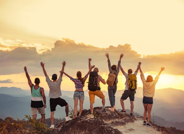 Group of happy friends stands with raised arms at sunset mountain and enjoys sunset