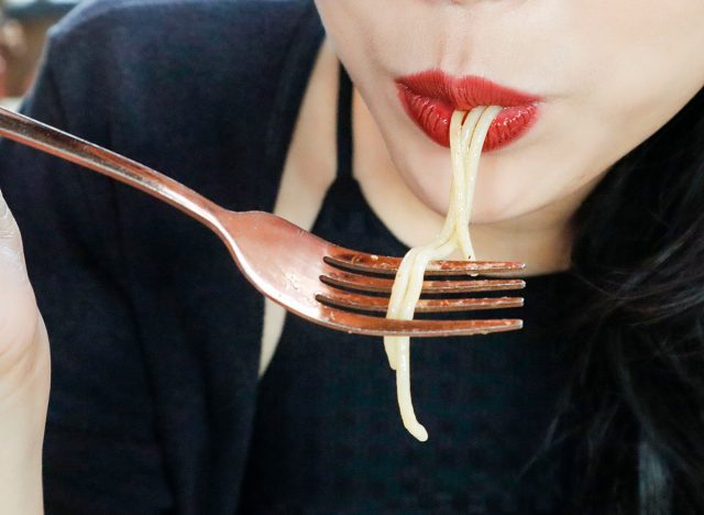 Close up of woman's mouth eating spaghetti by using her fork. Focus of a girl wearing red lipstick consuming her food.