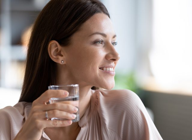 Head shot close up young dreamy woman holding glass of fresh pure water, looking aside. Happy lady visualizing future, planning workday, enjoying morning healthy habit at home, healthcare concept.