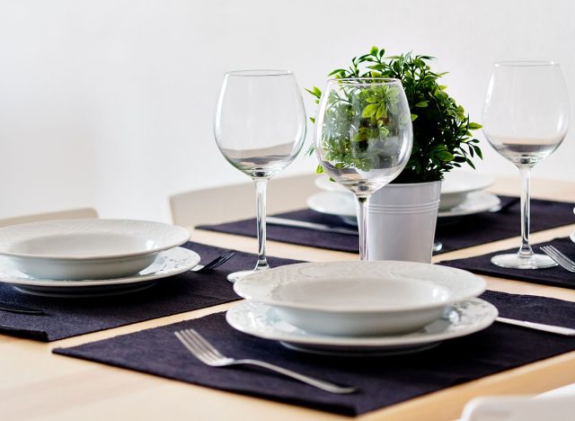 Close up new white porcelain tableware plates on purple place mats and empty wine glasses ready for dinner. Table settings wait for guests at home or restaurant, artificial potted plant for decoration