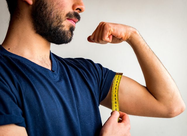 Bearded thin man measuring biceps, muscles of his left arm with a yellow tape measure. He's calm, serious, quiet. Wearing blue t-shirt. White background.