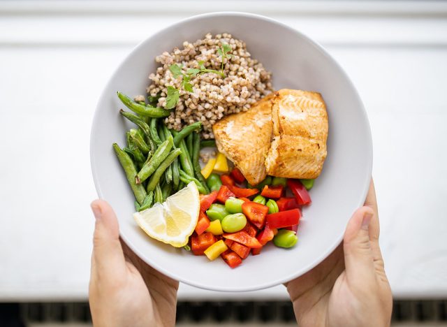 Hands holding salmon and buckwheat dish with green beans, broad beans, and tomato slices. Nutritious dish with vegetables and fish from above. Healthy balanced diet