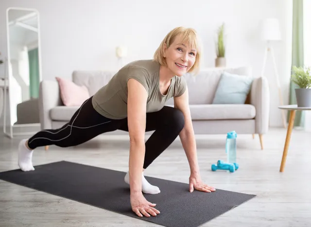 Positive athletic senior woman doing runner's lunge yoga pose on home workout, copy space. Sporty mature lady stretching her legs, exercising in living room. Domestic training during covid lockdown