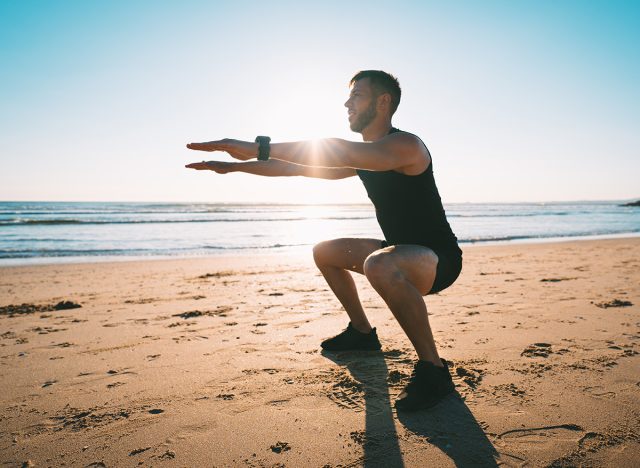 Young sporty man squatting or exercising on the beach during sunset. Athletic man doing fitness workout or training outdoors. Sports and healthy lifestyle