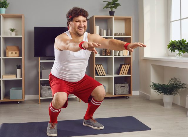 Training muscles and doing physical weight loss exercise to get fit. Funny chubby man having fitness workout at home. Happy fat guy in retro sportswear squatting standing on sports mat in living-room