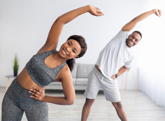 Millennial black couple doing lateral flexion exercise, working out together at home during coronavirus quarantine. Pretty young lady and her boyfriend making side bending pose, panorama