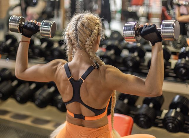 Fitness woman in sportswear trains shoulder muscles, doing dumbbell press while sitting on bench in gym. Back view