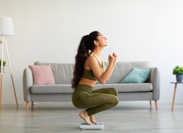 Full length of excited Indian woman sitting on scales at home, overjoyed with success of her slimming diet, side view. Emotional Asian lady achieving her weight loss goal, making YES gesture