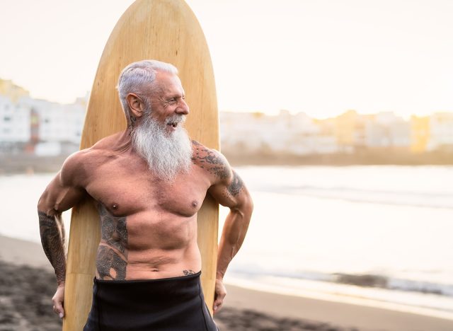 Senior male having fun surfing during sunset time - Fit retired man training with surfboard on the beach - Elderly healthy people lifestyle and extreme sport concept