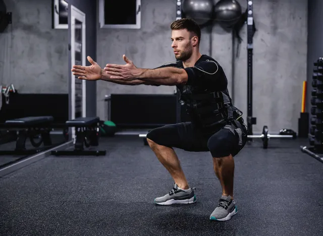 Leg burning exercise, using new EMS technology. A young attractive man in EMS clothes in the gym doing squats with his arms outstretched. Electrical muscle stimulation, strong movement