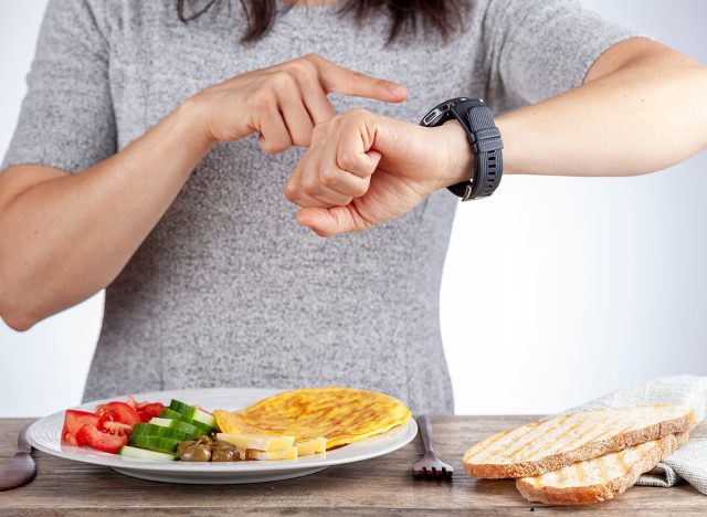 Intermittent fasting concept with a woman sitting hungry in front of food and looking at her watch to make sure she breaks fast on the correct time. A dietary modification for healthy lifestyle.