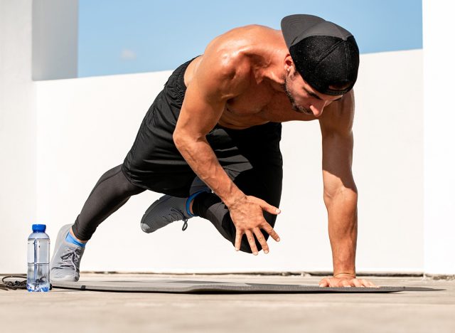 Shirtless athletic man doing high plank knee touch workout on rooftop floor, home open air exercise concept, selective focus