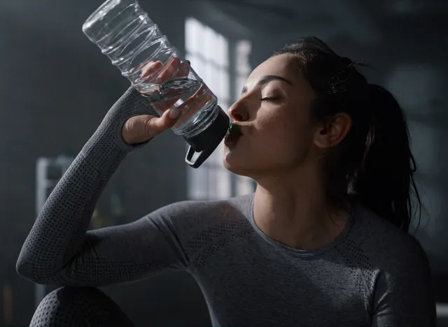 Exhausted woman in sportswear sitting box at fitness training in dark gym. Fit girl drinking water sports bottle at cardio workout. Tired athlete resting at sport club. Female bodybuilder making break