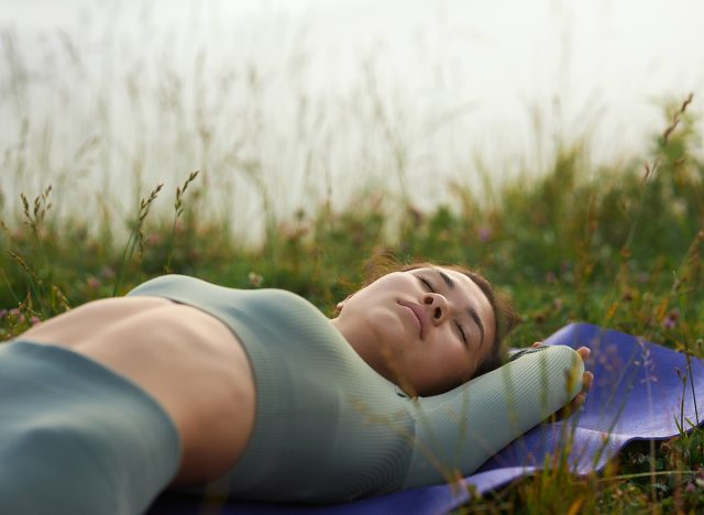 Side view of female in green wear sleeping outdoors after doing yoga exercises. Crop of woman relaxing peacefully after yoga exercising among grass holding hands behind head. Concept of calmness.