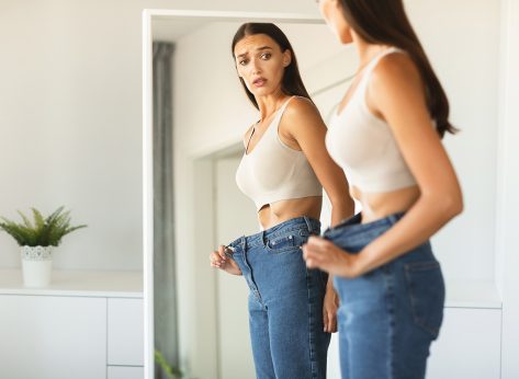 Shocked young woman wearing too big loose jeans after successful weight loss, posing and looking at her reflection in mirror with open mouth, copy space