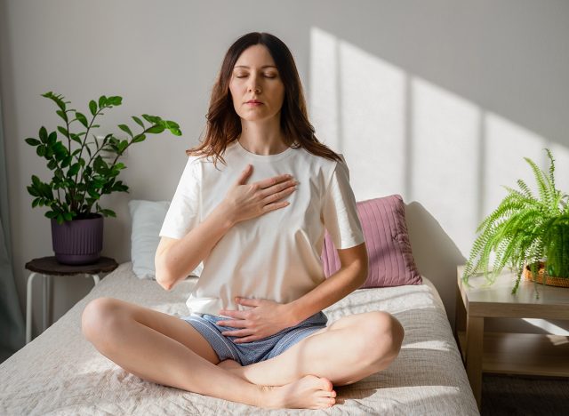 Pretty female sitting on bed in lotus posture with hands on her chest and belly doing pranayama techniques. Doing yoga at home. Self care and positive emotions.