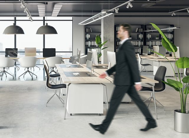 Front view of blank white isolated poster on a light grey wall at the entrance to modern loft office interior with concrete floor and walking businessman on window city view background. Mockup