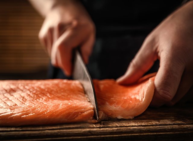 Chief hands cut salmon fillet with knife on wooden table at kitchen. Man cooking red omega fish with lemons for healthy nutrition diet