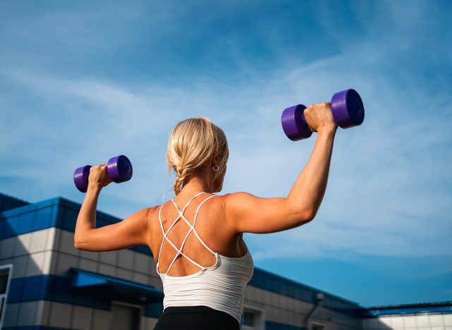 Beautiful blonde middle age woman doing weights exercises with dumbbells outdoors, sunny summer evening. Healthy active lifestyle, body conscious