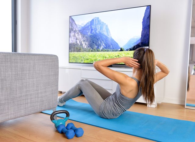 Home workout - woman exercising in front of a flat screen watching a fitness program or exercising during a TV show lying on a yoga mat in front of the sofa in the living room of a house or apartment.