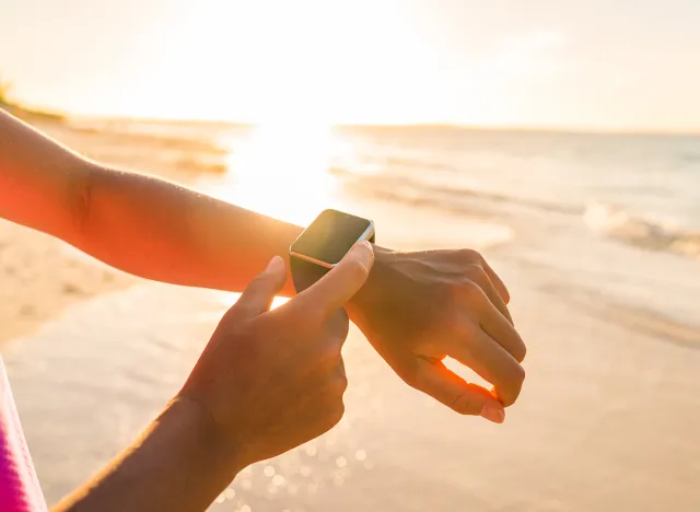 Smart watch woman using smartwatch touching button and touchscreen on active sports activity or morning jogging during beach sunrise or sunset. Closeup of hands and wrist with smart watch screen.