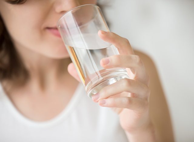 Happy beautiful young woman drinking water. Smiling caucasian female model holding transparent glass in her hand. Closeup. Focus on the arm