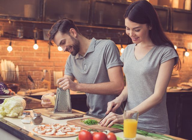 Pizza time. Young beautiful couple in kitchen. Family of two preparing food. Couple making delicious pizza. Man grating cheese. Nice loft interior with light bulbs