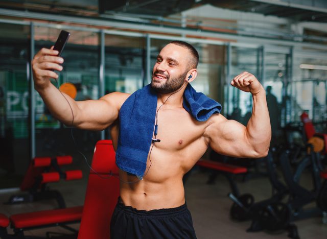 A gorgeous and muscular man flexing and showing off his arm muscles on a blurred gym background. A manly bodybuilder with blue towel, headphones and smartphone taking selfies of his hot, fit body.