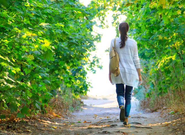 The girl walks along the path in the woods to the light in a white jacket and jeans.