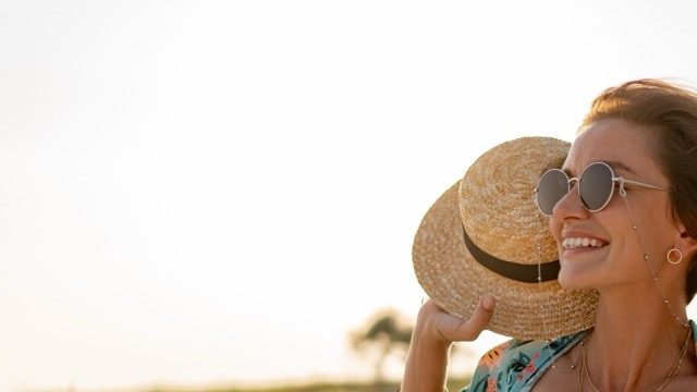 Pretty attractive slim smiling woman on sunny beach in summer style fashion trend outfit happy, freedom, wearing white top, jeans and colorful printed tunic boho style chic and straw hat.
