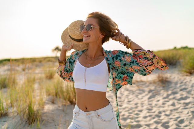 Pretty attractive slim smiling woman on sunny beach in summer style fashion trend outfit happy, freedom, wearing white top, jeans and colorful printed tunic boho style chic and straw hat.