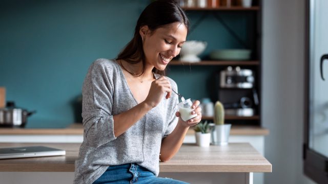 Shot of smiling young woman eating yogurt while sitting on stool in the kitchen at home.