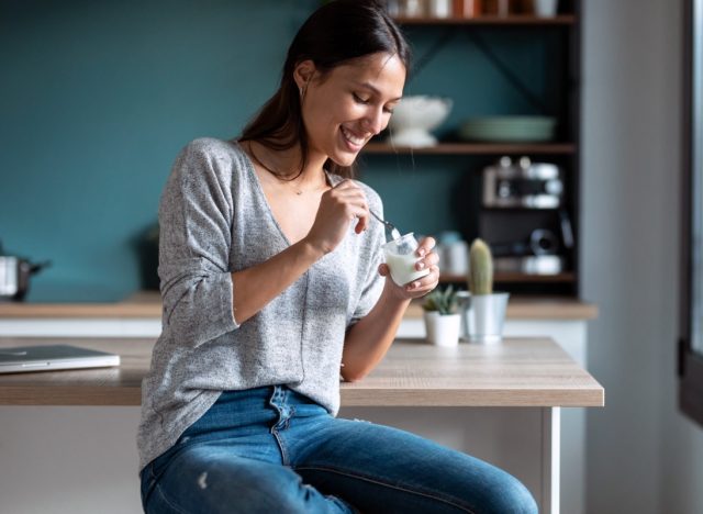 Shot of smiling young woman eating yogurt while sitting on stool in the kitchen at home.