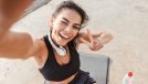 Cheerful young sportswoman resting after workout at the beach, taking a selfie, drinking water, sitting on a fitness mat