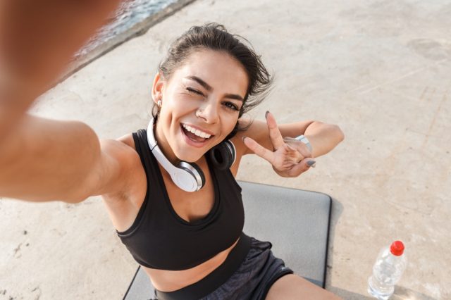 Cheerful young sportswoman resting after workout at the beach, taking a selfie, drinking water, sitting on a fitness mat