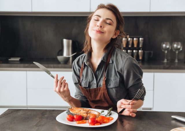 Image of young woman sitting in kitchen while eating fish and tomatoes.