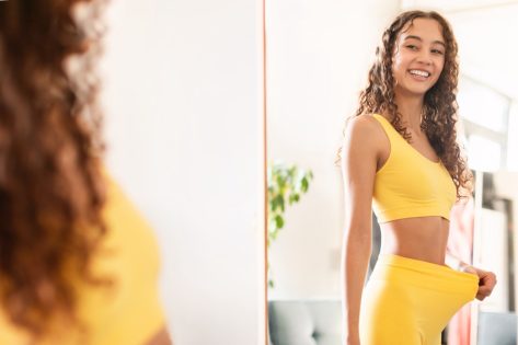 Happy fit woman looking at reflection in mirror and pulling fitness pants waist showing result of dieting and slimming, motivated after workout training at home.