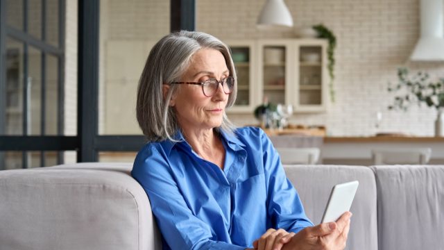 Serious mature middle age senior woman at home on couch holding mobile cellphone.