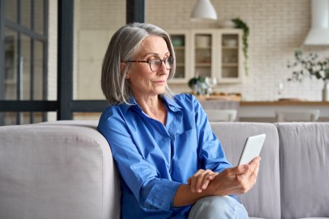 Serious mature middle age senior woman at home on couch holding mobile cellphone.