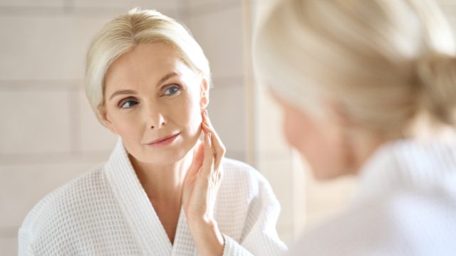 Headshot of mid age adult 50 years old blonde woman standing in bathroom after shower touching face, looking at reflection in mirror doing morning beauty routine.